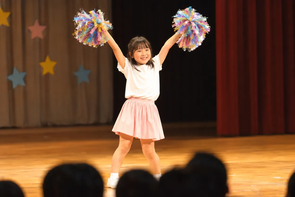 発表会のステージで、手作りのポンポンを持って腕を高く上げて踊るチアの女の子